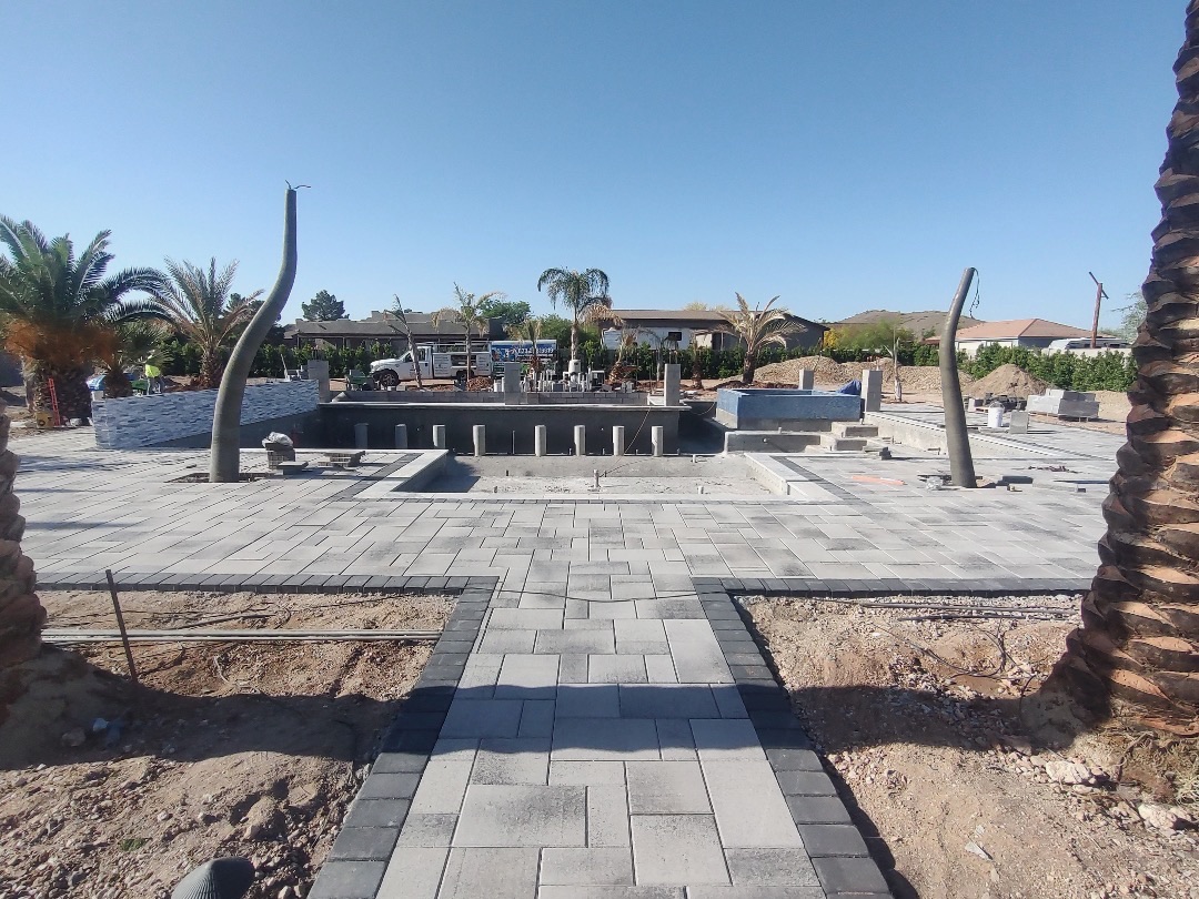 Aurora Estate — wide paver field set along the main axis with the pool shell mid-build, stone columns staged, and a paver walkway leading to the house