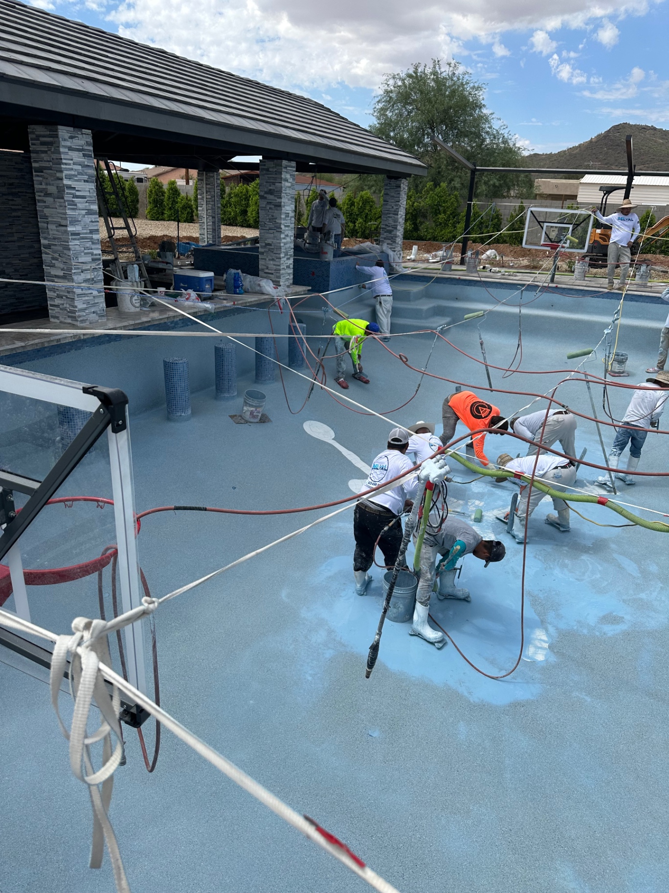 Aurora Estate — full pebble-finish crew hand-troweling the pool interior with the ramada and basketball hoop in the background