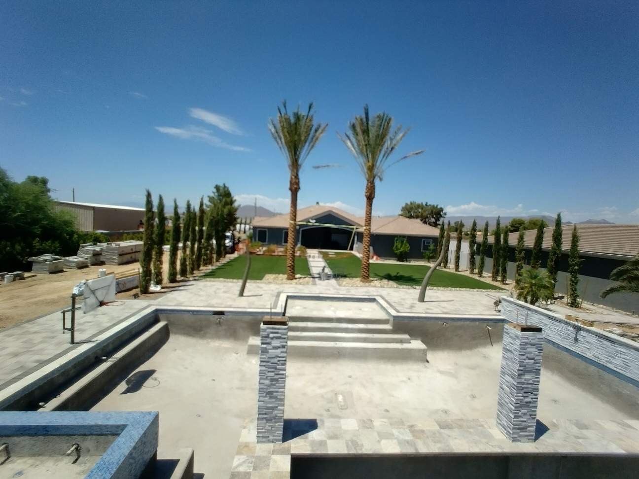 Aurora Estate — wide elevated view of the finished pool shell with stacked-stone columns, sun shelf, and the house framed by twin date palms