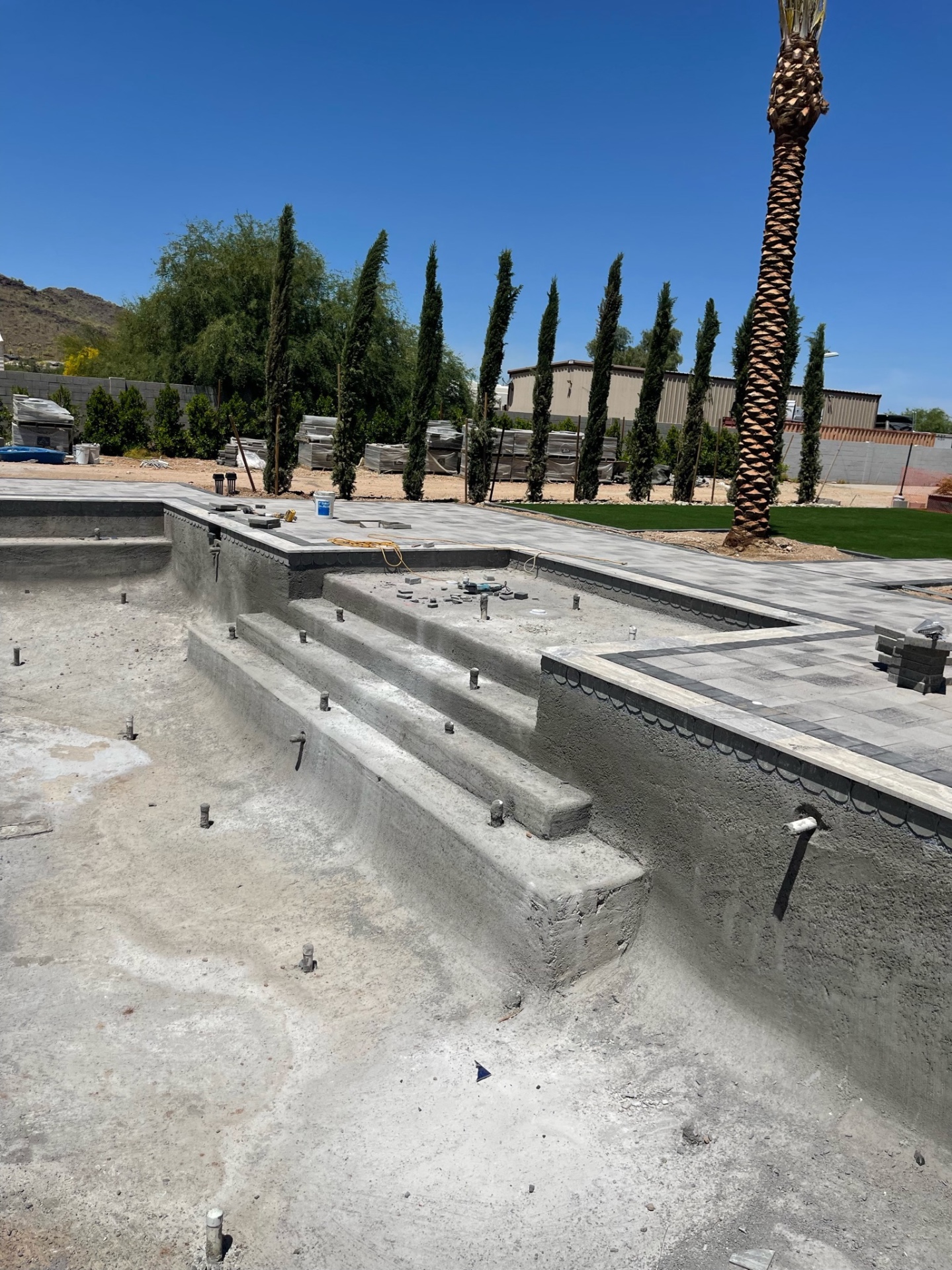 Aurora Estate — pool steps and shallow shelf inside the empty shell with charcoal paver banding and deck geometry already locked around the perimeter