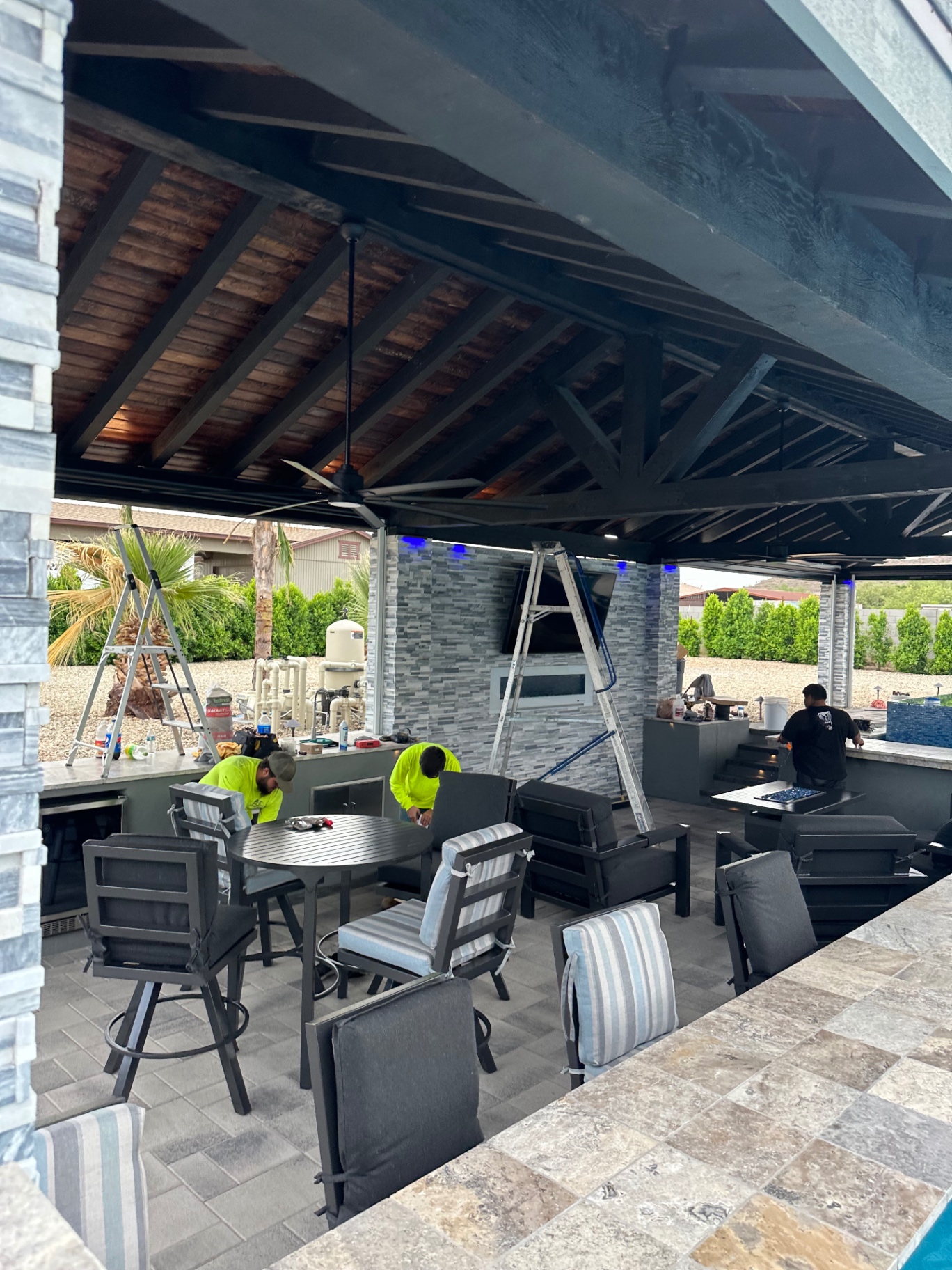 Aurora Estate — ramada interior nearing completion with outdoor kitchen, TV install in progress, fan and lounge seating staged