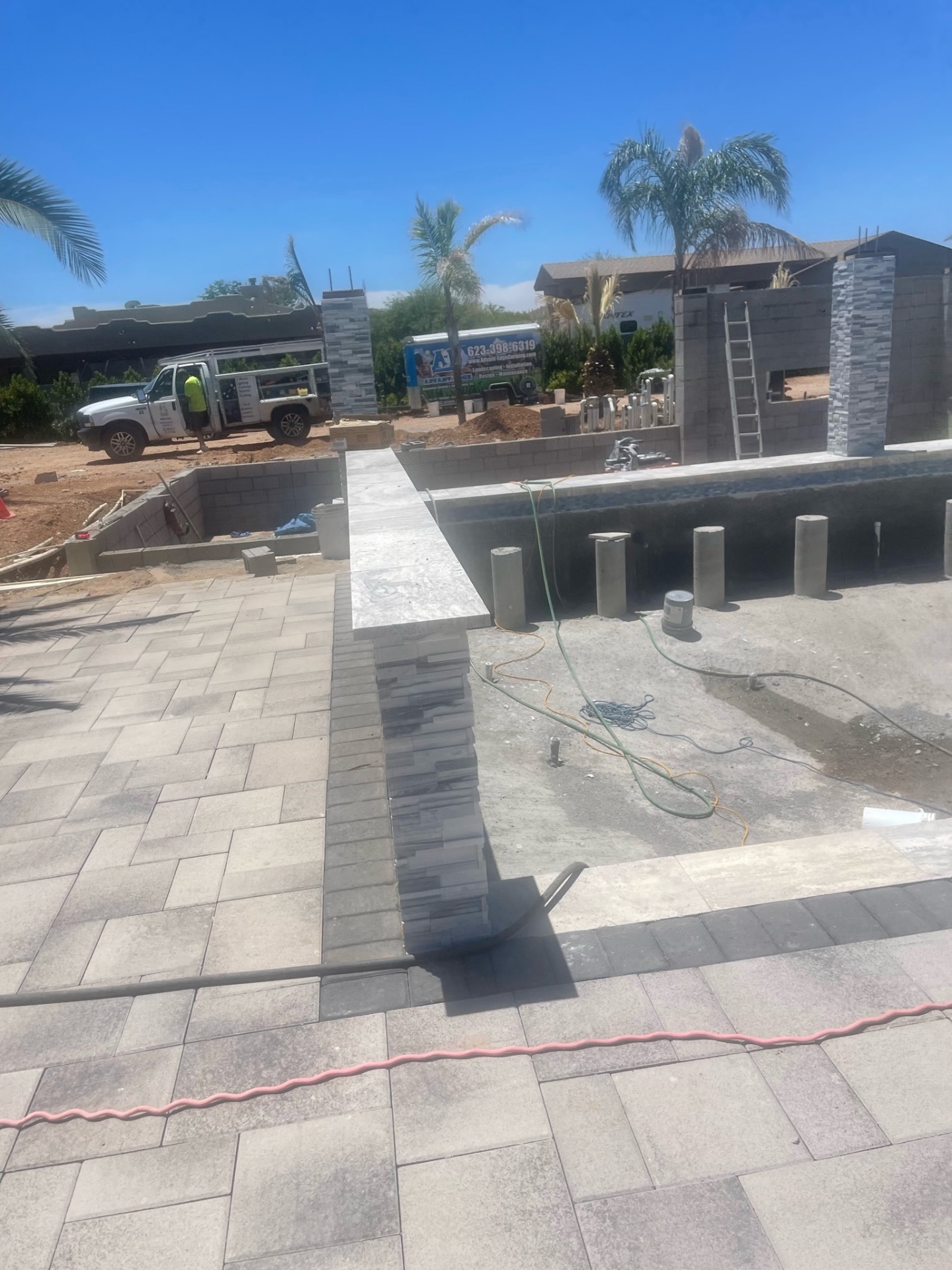 Aurora Estate — finished paver walkway crossing a stacked-stone column, pool shell visible alongside and outdoor kitchen block walls in the background